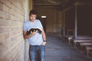 Man leaning against brick wall at a college reading a book
